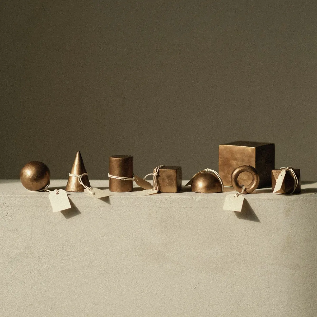 Seven bronze solids in a row on a pale plaster ledge, most tied with cord to blank paper tags: a sphere, cone, cylinder, cube, half-dome, ring and faceted block, with one larger untagged cube behind them against a dark olive wall in warm side light